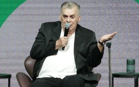 De blazer e calça preta, Walcyr Carrasco está sentado em uma cadeira, falando com um microfone